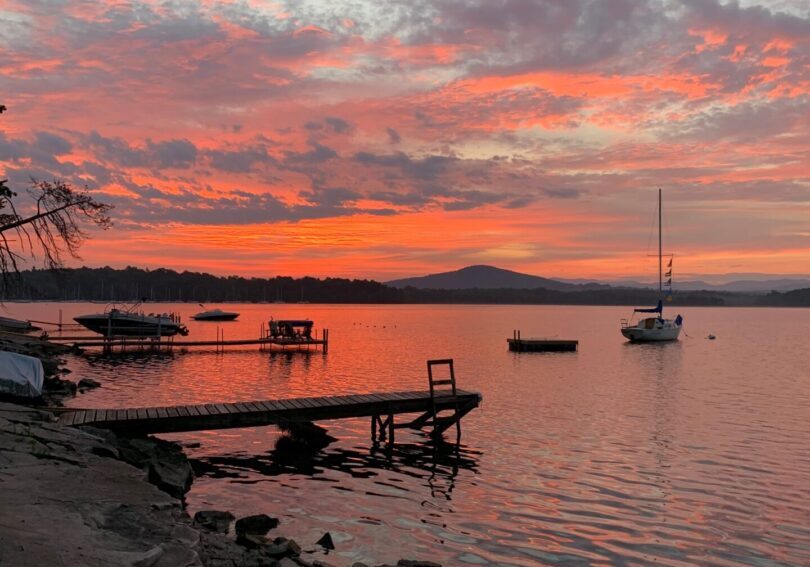 Sunset over a calm lake with boats and rocky shore.