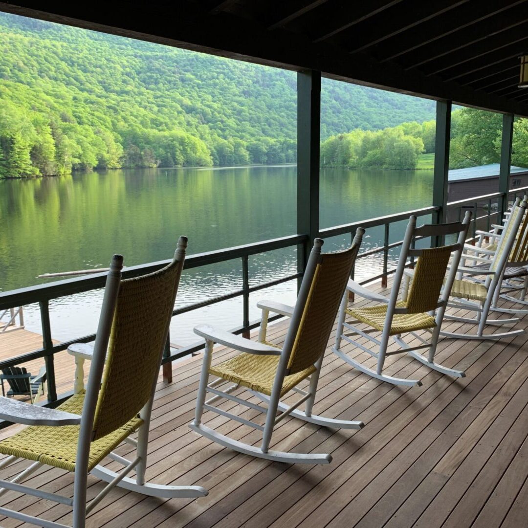 Rocking chairs lined up on a porch overlooking a calm river and green hills.