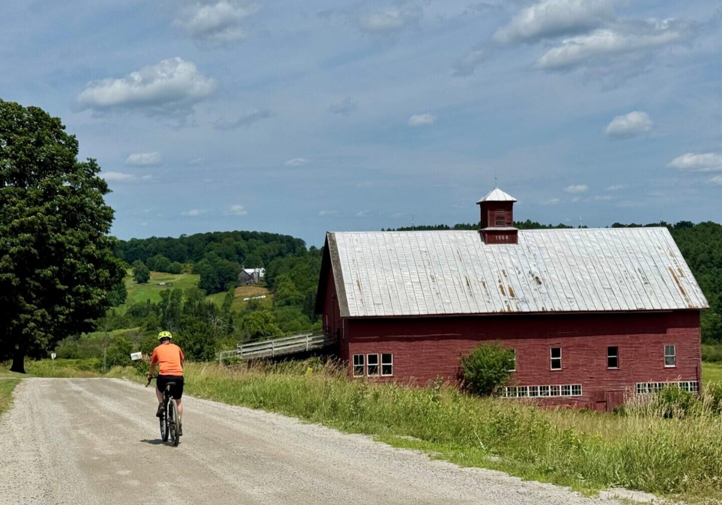 A person cycling on a rural dirt road past a red barn.